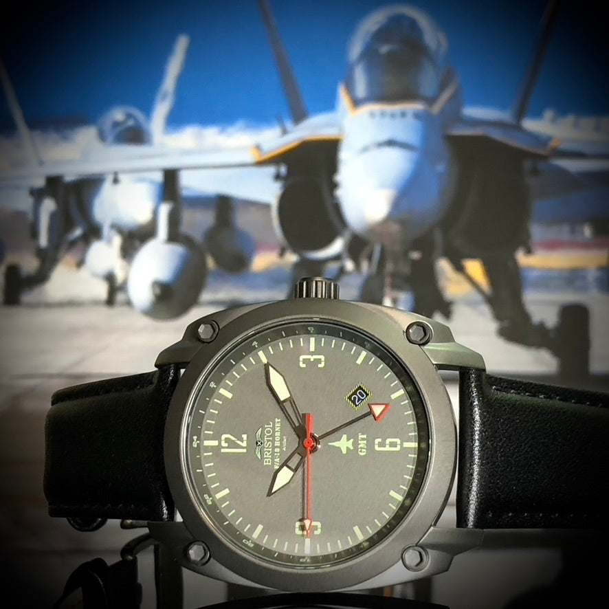 F-18 Hornet tribute watch with gunmetal finish, gray dial, black leather band, and Swiss quartz movement in front of F-18 Hornet jets.