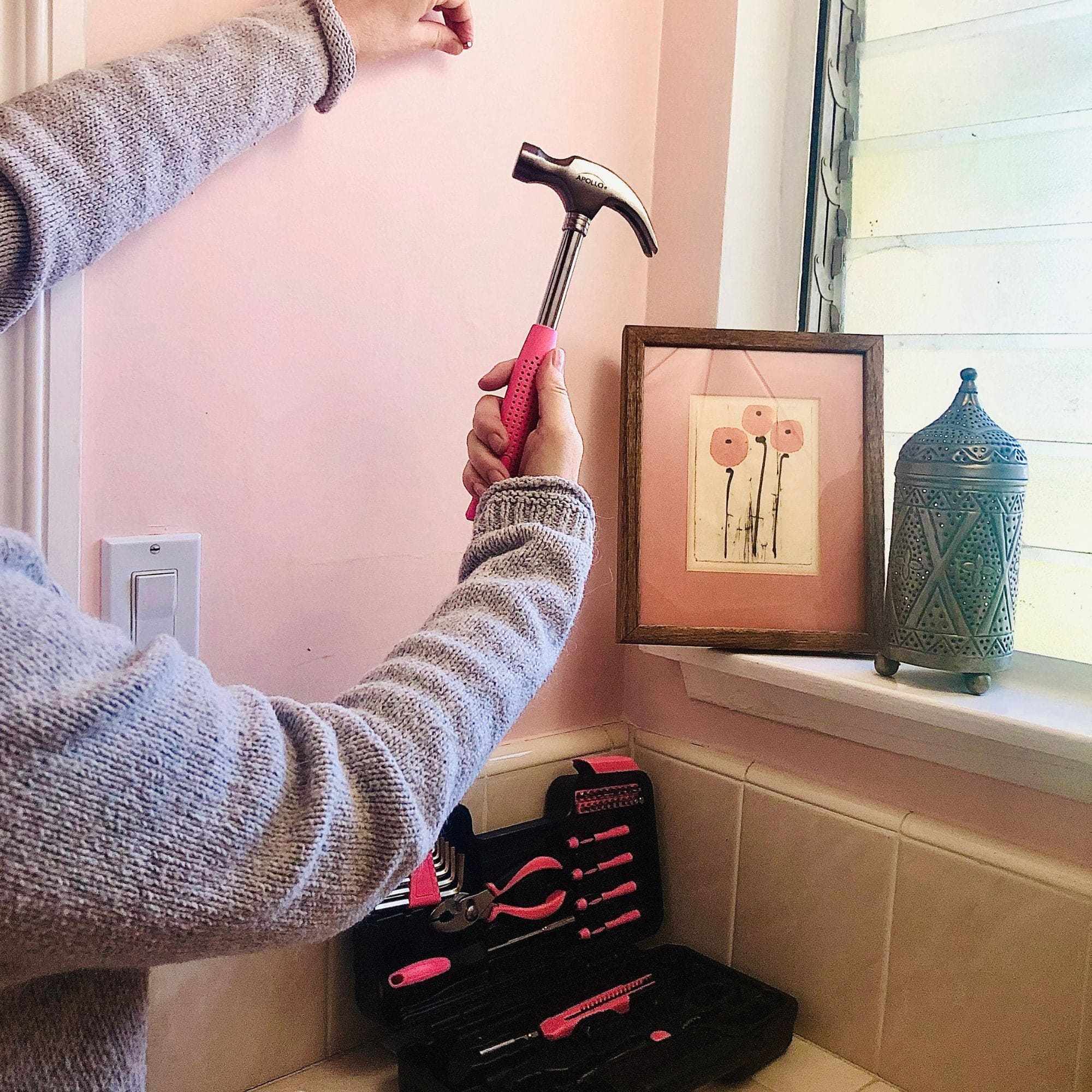 Person using pink hammer from 39 Piece General Tool Set for home repair near framed wall art and decorative jar