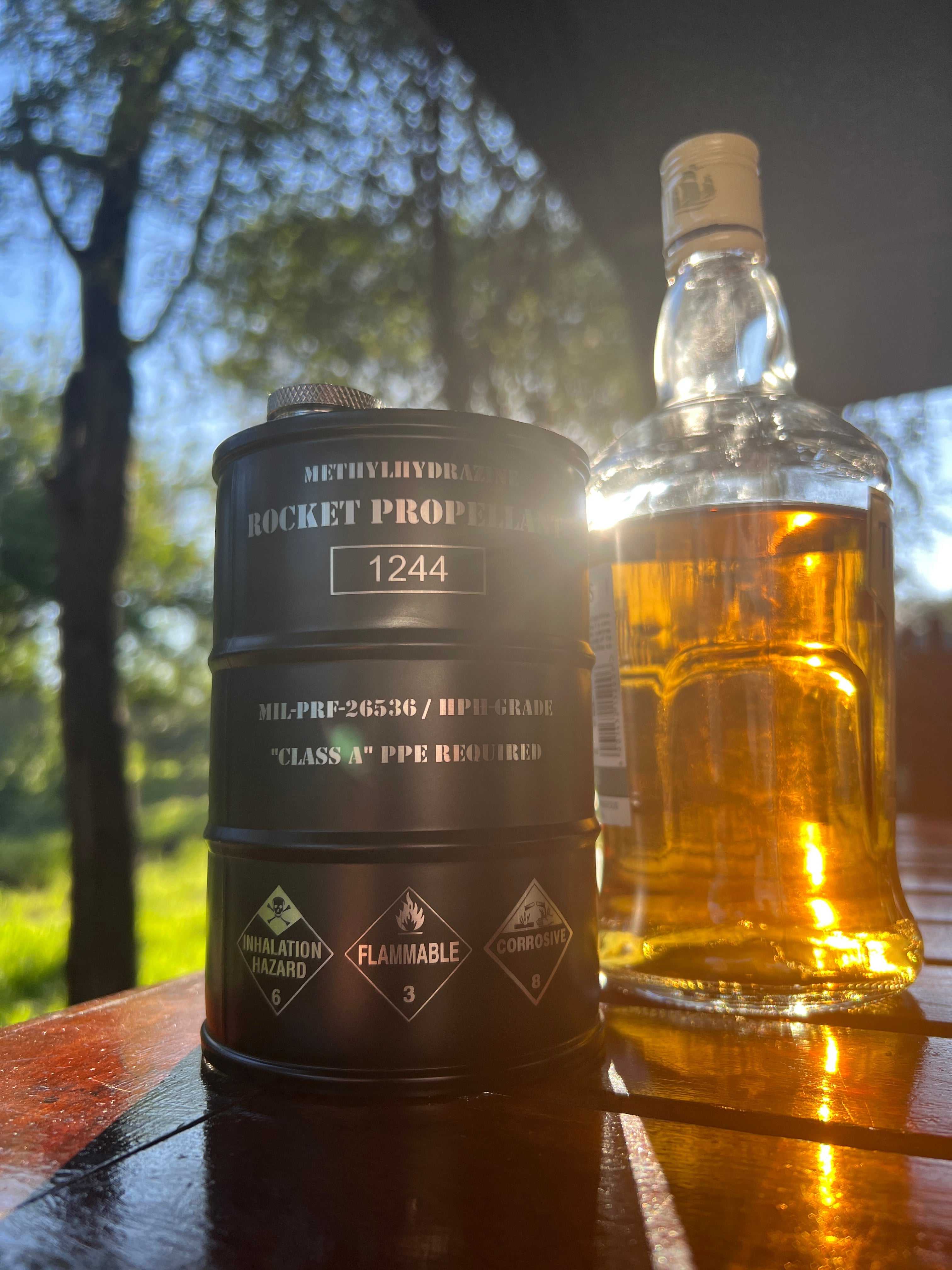 Hydrazine Barrel Flask shaped like a NASA fuel drum with hazard symbols and black matte finish next to a bottle of amber liquid outdoors