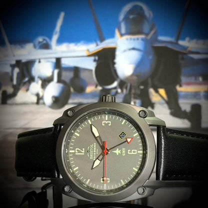 F-18 Hornet tribute watch with gunmetal finish, gray dial, black leather band, and Swiss quartz movement in front of F-18 Hornet jets.