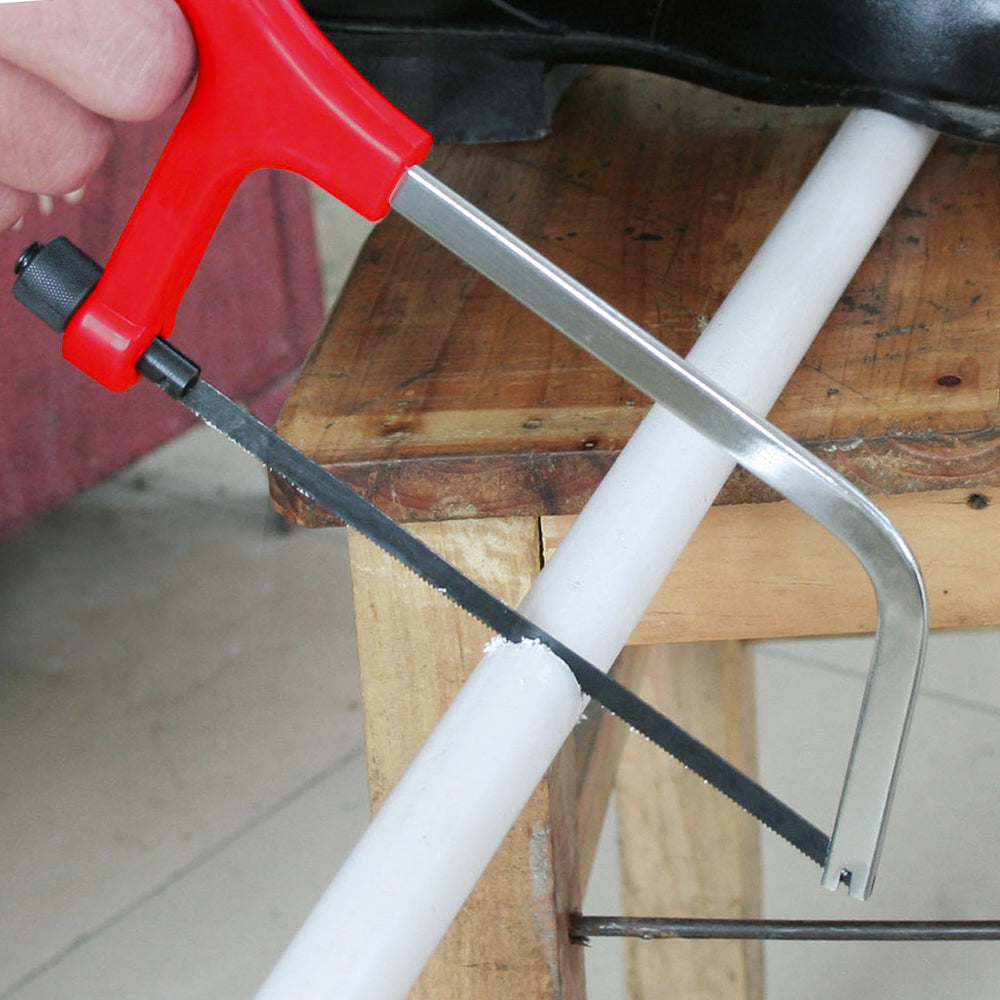 Person using a red-handled hacksaw to cut a white PVC pipe on a wooden surface, part of a household tool kit.