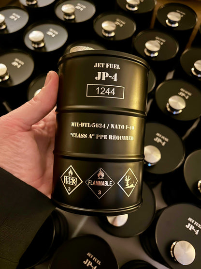 Hand holding a black JP-4 Jet Fuel Flask shaped like a fuel barrel with hazard symbols and industrial markings, surrounded by similar flasks.