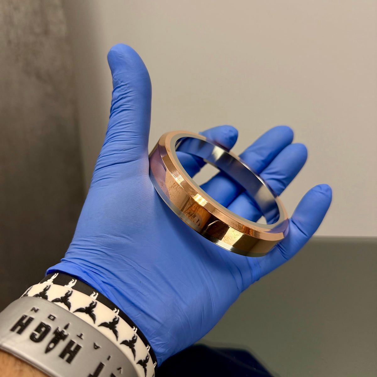 Hand wearing a blue glove holding a metallic bracelet against a neutral background