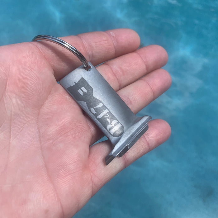 Hand holding a silver keychain with 'Belay' branding against a blue water background