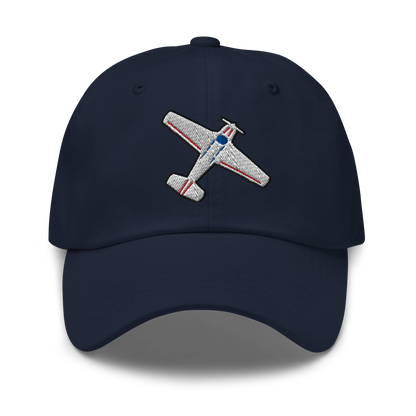A navy blue baseball cap featuring an embroidered airplane motif on the front panel. The airplane design is detailed with a textured silver body, red stripes, and a blue nose section.