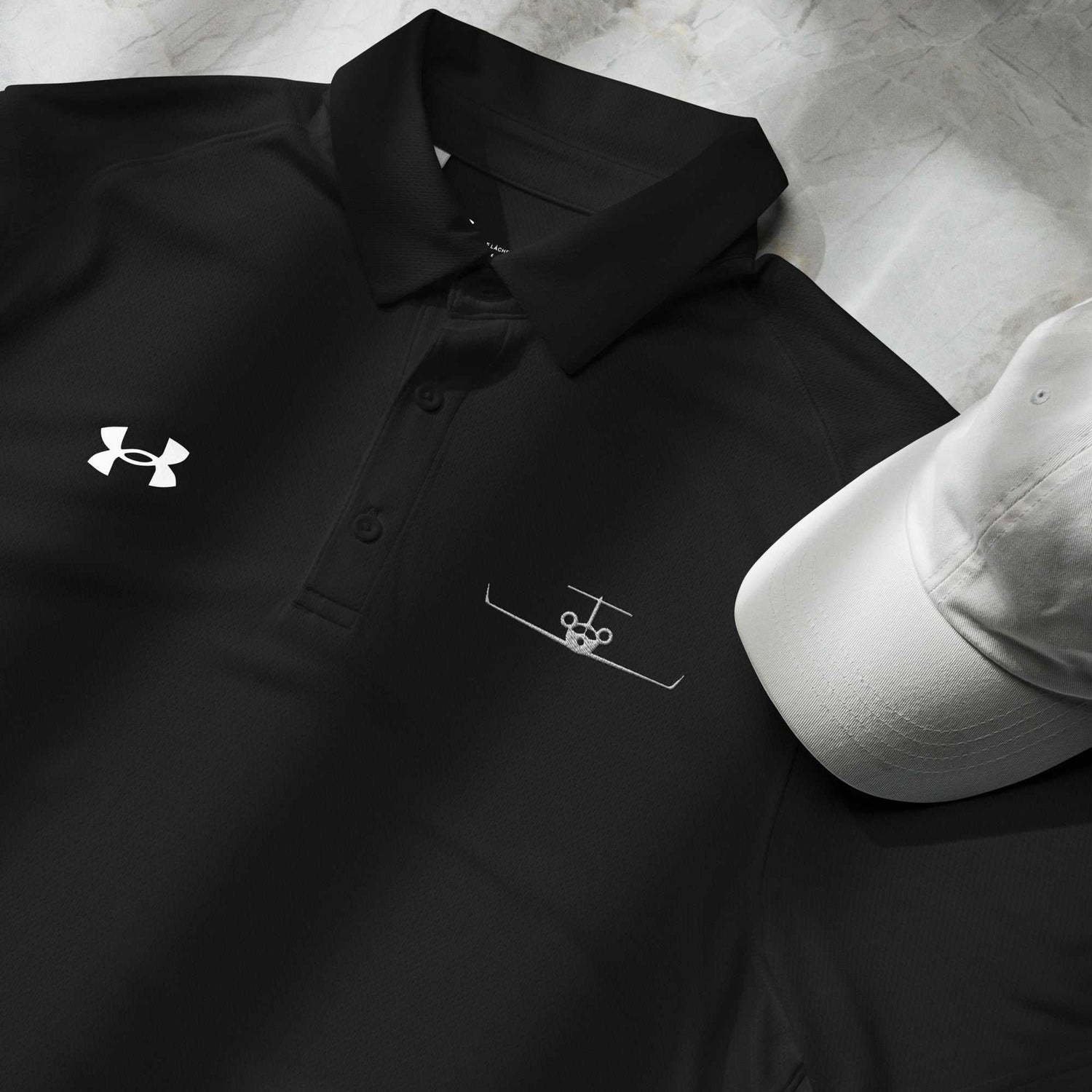 Black pilot polo shirt with embroidered private jet logo and Under Armour logo next to a white cap