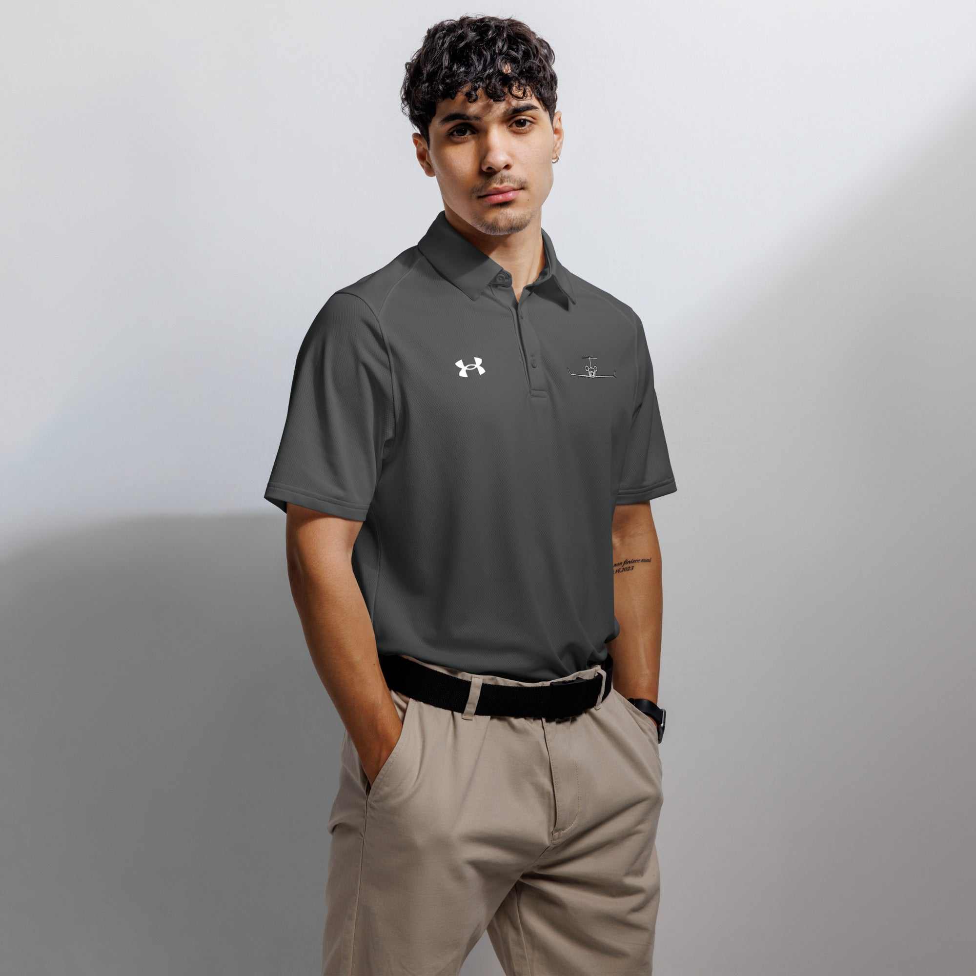 Man wearing black pilot polo shirt with embroidered private jet logo and Under Armour logo, paired with beige pants