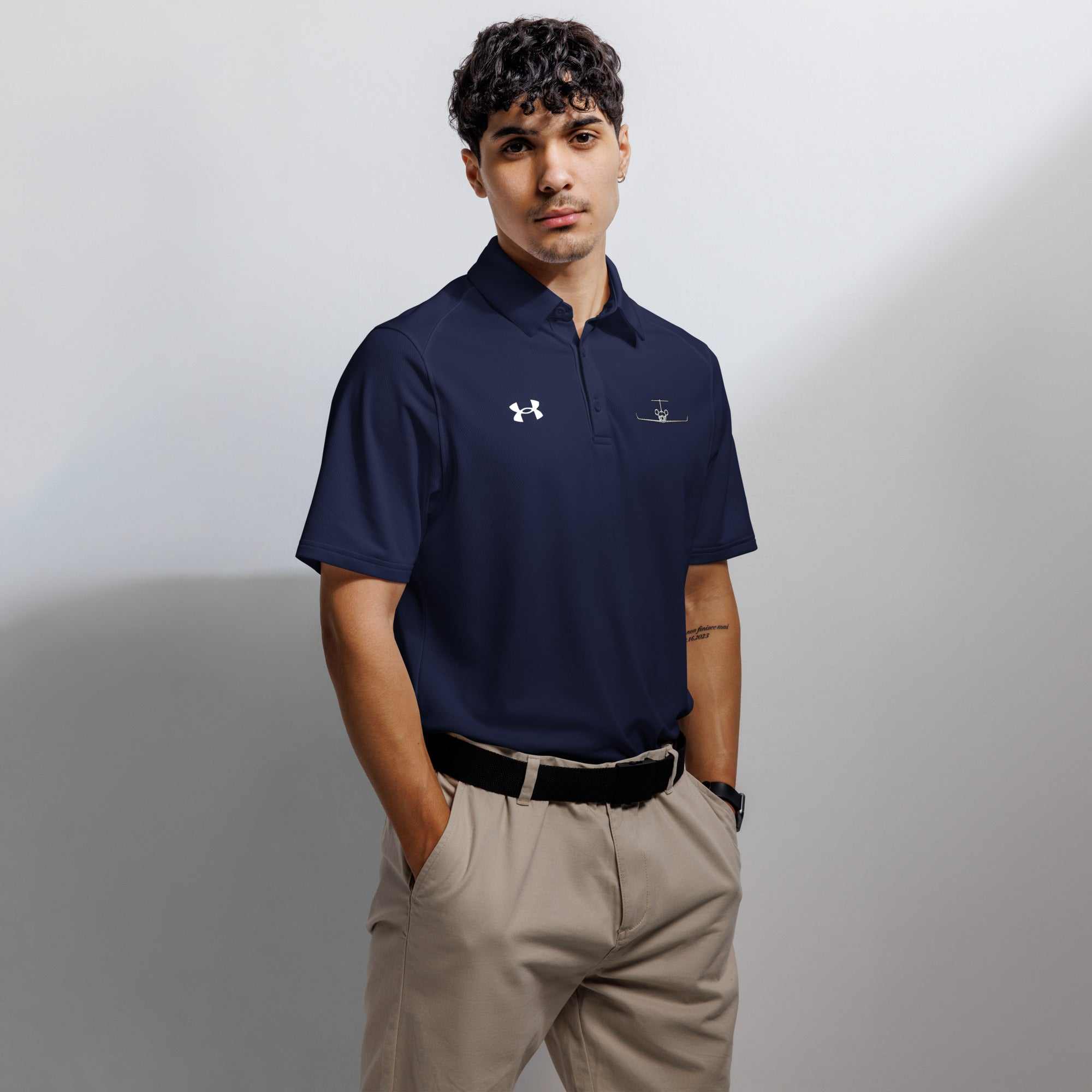 Man wearing navy pilot polo shirt with embroidered private jet logo and Under Armour logo, paired with beige pants, against gray background
